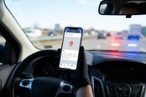 Vue du poste de conduite d'une voiture. Un conducteur tient un smartphone affichant un écran "LIVE" avec des gyrophares de police devant.