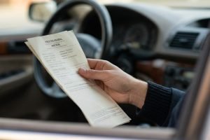 excès de vitesse sans radar Peut-on être sanctionné sans preuve d'excès de vitesse ? Main tenant un procès-verbal d'amende dans une voiture. Le volant et le tableau de bord sont flous en arrière-plan.