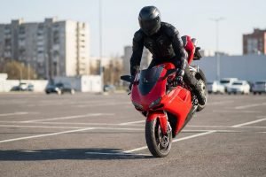 wheeling en parking public ou rodéo motorisé. Un pilote entièrement équipé fait un wheelie sur une moto de sport rouge dans un grand parking asphalté. Bâtiments en arrière-plan.